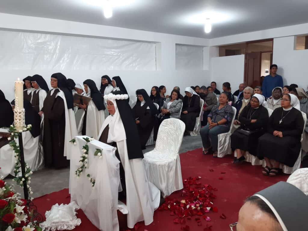 A group of nuns in black and white habits are gathered in a decorated room. Some are seated, others are standing, with flower petals on the red carpet and a large candle nearby. Several attendees are watching the ceremony.