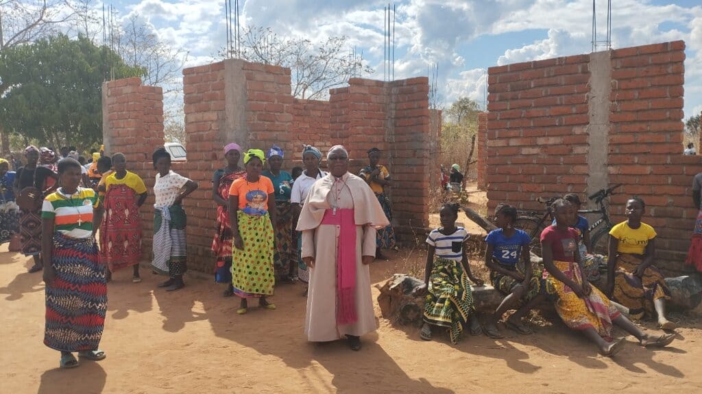 Un grupo de mujeres y niñas, posan frente a un edificio de ladrillo a medio construir bajo un cielo nublado en una zona rural, junto al sacerdote de su parroquia