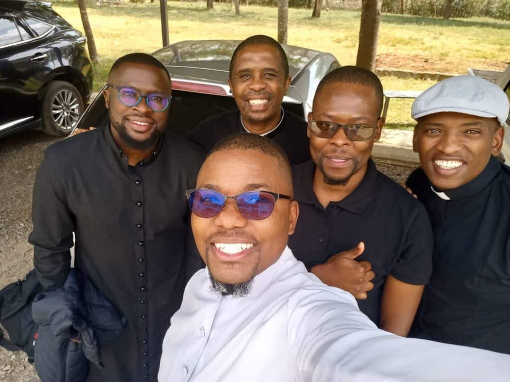 Cinco seminaristas sonrientes posan al aire libre para una selfie. Visten ropa oscura y clara, y algunos llevan cuellos clericales. Al fondo se ven árboles, coches aparcados y césped.