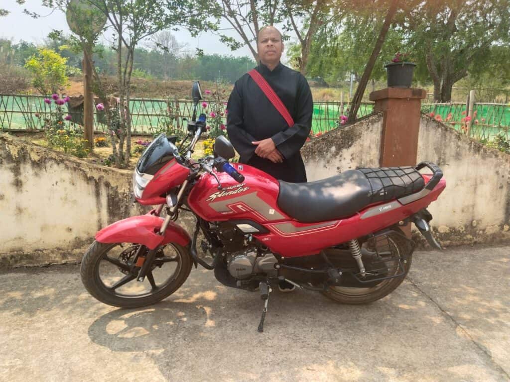 A man in a black robe with a red sash stands outdoors beside a red motorcycle, with a garden and trees in the background.