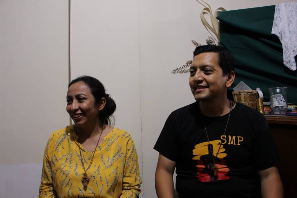 A woman in a yellow patterned top and a man in a black SMP shirt are sitting and smiling indoors, with a decorative cloth and baskets in the background.