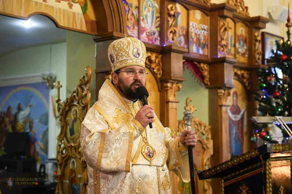 A bearded priest in ornate gold vestments and a mitre speaks into a microphone and holds a staff inside a richly decorated church with religious icons and a Christmas tree in the background.